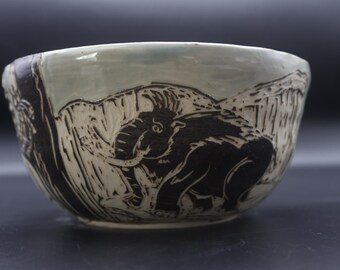 May include: A ceramic bowl with a hand-carved design featuring a black elephant and mountain landscape. The bowl has a light blue interior and a white exterior with black detailing. The bowl is a unique piece of art.