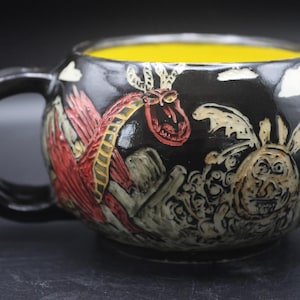May include: Black ceramic mug with a yellow interior. The exterior features a detailed relief design of a red dragon, a creature, and clouds. The handle is black. The mug is a unique piece of art.