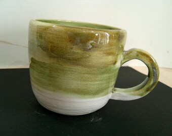 May include: Handmade ceramic mug with a unique, organic design. The mug features a rounded shape with a handle and is painted with shades of green, brown, and white. The mug is suitable for coffee, tea, or other beverages.