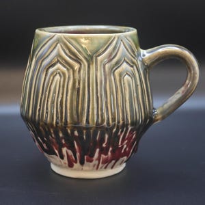 May include: A handmade ceramic mug with a unique textured design. The mug features a green and brown glaze with red accents near the base. It has a rounded body, a handle, and a white base.