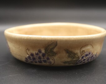 May include: A beige ceramic bowl with a rounded shape. The bowl is decorated with a painted design of purple grapes and green leaves. The bowl has a smooth, matte finish and is suitable for serving food.
