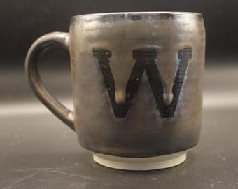 May include: A handmade ceramic mug with a dark, textured glaze. The mug features a large, black letter "W" on the front. The handle is curved, and the base is a lighter color. This mug is suitable for coffee, tea, or other beverages.