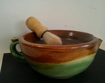 May include: A shaving set featuring a ceramic bowl with a handle and a shaving brush. The bowl is brown, orange, and green. The brush has a wooden handle and light-colored bristles. The set is designed for grooming.