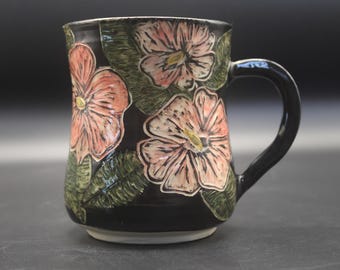 May include: A ceramic mug with a glossy black glaze featuring hand-painted pink flowers and textured green leaves. The mug has a curved handle on the right side and a white base. The floral design is embossed with visible brushstrokes.