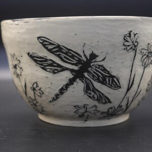 May include: A white ceramic bowl with a textured, uneven surface featuring a black ink-style illustration of a large dragonfly centered among several small wildflowers. The bowl has a rounded shape and a smooth, circular base.