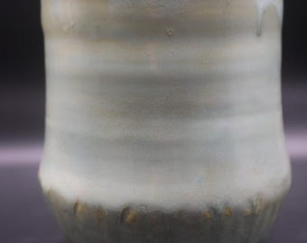 May include: A handmade ceramic tumbler with a light green and beige glaze. The cup has a rounded base and a slightly flared rim. The bottom edge features small, textured details. The overall aesthetic is rustic and organic.