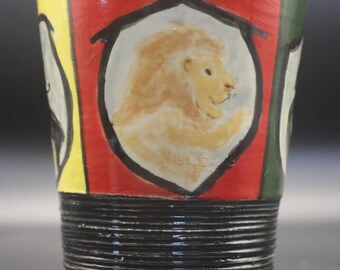 May include: A ceramic tumbler featuring a hand-painted lion inside a shield emblem on a red background. The upper portion displays vertical panels of yellow, red, and green, while the lower section has a dark, ribbed, horizontal texture.