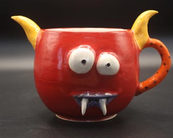May include: A bright red ceramic mug shaped like a monster with two yellow horns, two large white eyes with black pupils, a blue mouth, and two white fangs. The handle is orange with small black spots, and the mug has a glossy finish throughout.