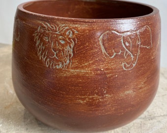 May include: A brown ceramic bowl with a textured surface. The bowl has a raised design of a lion and two elephants around the rim.