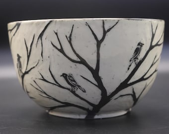 May include: A ceramic bowl with an off-white base color featuring a hand-painted black design of bare tree branches and small birds perched on the limbs. The bowl has a rounded shape and a smooth, glossy finish across the exterior surface.