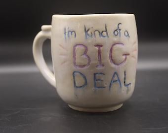 May include: Cream-colored ceramic mug with the phrase "I'm kind of a BIG DEAL" in blue, purple, and pink lettering. The mug has a curved handle and a slightly flared rim. A fun, handmade coffee mug.