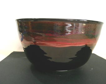 May include: A handmade ceramic bowl with a glossy finish. The bowl transitions from a deep black base to a band of red, brown, and green hues near the top. The bowl is likely used for serving or display.