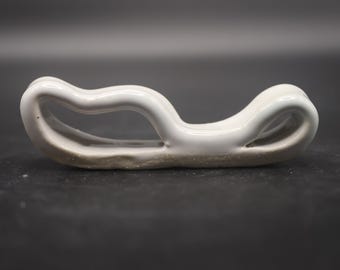 May include: A white ceramic object with a curved, open design. The piece has a smooth, glossy finish and rests on a textured, light brown base. The shape is reminiscent of a stylized wave or loop.