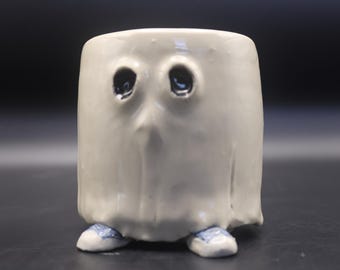 May include: A cylindrical ceramic mug with a glossy off-white glaze, shaped like a ghost with two dark, recessed eye sockets. The base of the mug features two small, light blue painted feet that resemble sneakers, protruding from the bottom edge.