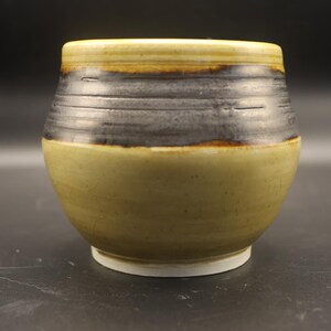 May include: A handcrafted ceramic bowl with a rounded form. The bowl has a two-tone design, with a light olive green base and a dark grey band around the middle. The rim is a lighter shade of olive green.