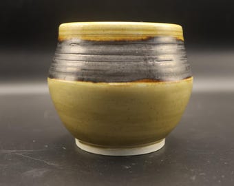 May include: A handmade ceramic bowl with a rounded shape. The bowl features a two-tone design, with a light olive green base and a dark gray band around the middle. The rim is a lighter shade of olive green.