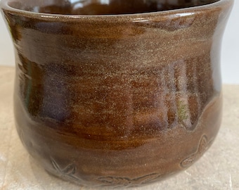 May include: A brown ceramic planter with a glossy finish and a subtle, textured design. The planter features a slightly flared rim and a rounded base.