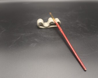 May include: A red-handled paintbrush rests on a small, off-white ceramic brush holder. The brush has a gold-colored bristle end. The items are set against a dark gray background. This image is suitable for art supplies.