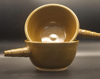 May include: Two stacked, handmade ceramic bowls in a warm, earthy brown glaze. Each bowl has a rounded shape with a small, spout-like handle. The bowls are set against a dark background.