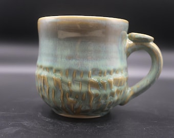 May include: A handmade ceramic mug with a textured, earthy design. The mug features a rounded body with a textured lower half in shades of brown and green. The upper portion and handle are a lighter green. The mug is approximately 4 inches tall.