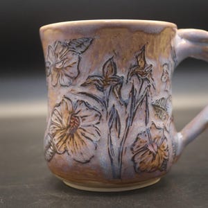 May include: A ceramic mug with a light purple and tan glaze featuring carved floral patterns. The mug has a rounded handle and a textured surface with dark outlines highlighting the embossed flowers and leaves across the exterior body.