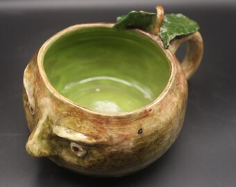 May include: A handcrafted ceramic mug with a unique apple design. The exterior features a textured, earthy brown and green glaze, while the interior is a vibrant green. The mug has a handle and a leaf detail, adding to its artistic appeal.