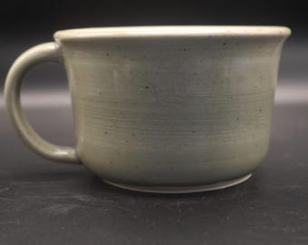 May include: A handmade, light green ceramic mug with a rounded body and a curved handle. The mug has a textured surface with visible horizontal lines and small dark speckles. The rim is slightly flared, and the mug sits on a white base.