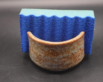 May include: A ceramic sponge holder with a semi-circular shape and a speckled tan and light blue glaze. The holder contains a rectangular blue sponge with a wavy, textured surface on one side, designed to fit securely within the curved ceramic.
