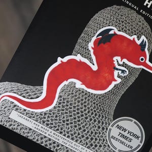 May include: A red worm-like sticker with black accents is placed on a book. The book has a black cover with the title "HEAR" visible. A "New York Times Bestseller" sticker is also present. The sticker is on top of a chainmail pattern.