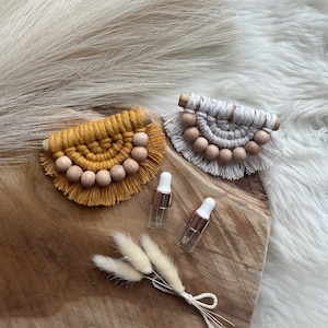 Può includere: Primo piano di due decorazioni da parete in macramè giallo senape e bianco, ciascuna con perline di legno. Sono presenti anche due piccoli flaconi di vetro con dettagli oro rosa e un mazzo di fiori secchi. Gli oggetti sono disposti su una superficie di legno.