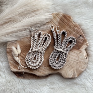 Puede incluir: Dos correas de estilo macramé con cierres plateados, una en beige y blanco, la otra en beige liso. Ambas están atadas con hilo y descansan sobre una tabla de madera con un fondo blanco y esponjoso. Flores secas decorativas atadas con hilo.