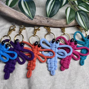 Op de afbeelding: Een verzameling kleurrijke sleutelhangers in de vorm van libellen. Elke sleutelhanger heeft een libellenontwerp gemaakt van geknoopt koord in verschillende kleuren, waaronder blauw, paars, oranje, roze en turquoise. De sleutelhangers hebben goudkleurige hardware en zwarte kraalaccenten.