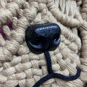May include: Close-up of a crocheted dog face with a black plastic nose and black yarn for the mouth.