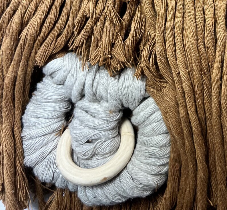 May include: Close-up of a light brown and gray macrame knot with a white wooden ring. The knot is made of thick, soft fibers and has a natural, rustic look.