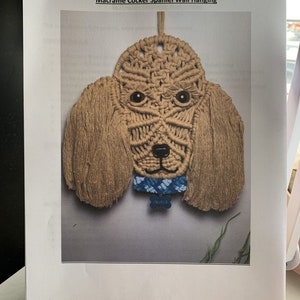 May include: A macrame wall hanging of a cocker spaniel dog with brown fur, blue eyes, and a blue and white collar with a tag that says "Charlie".
