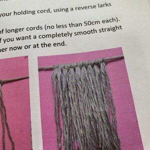 May include: A close-up of a gray macrame wall hanging with a knotted top and long strands hanging down. The macrame is on a pink background.