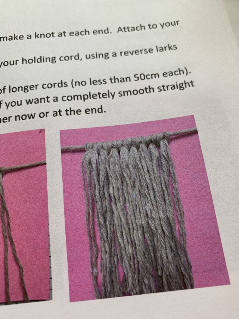 May include: A close-up of a gray macrame wall hanging with a knotted top and long strands hanging down. The macrame is on a pink background.