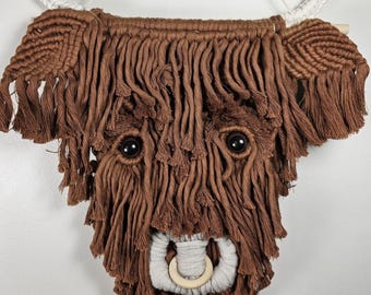 May include: Brown and white macrame wall hanging of a Highland cow head with black eyes.