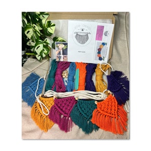 May include: A colourful macrame garland kit with instructions and supplies. The kit includes six different coloured cords, wooden beads, and a knot guide. The finished garland will have six triangular macrame pieces with tassels.