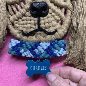 May include: A tan macrame dog wall hanging with a black nose and a blue and green macrame collar. The dog is wearing a blue bone-shaped tag that says "CHARLIE".