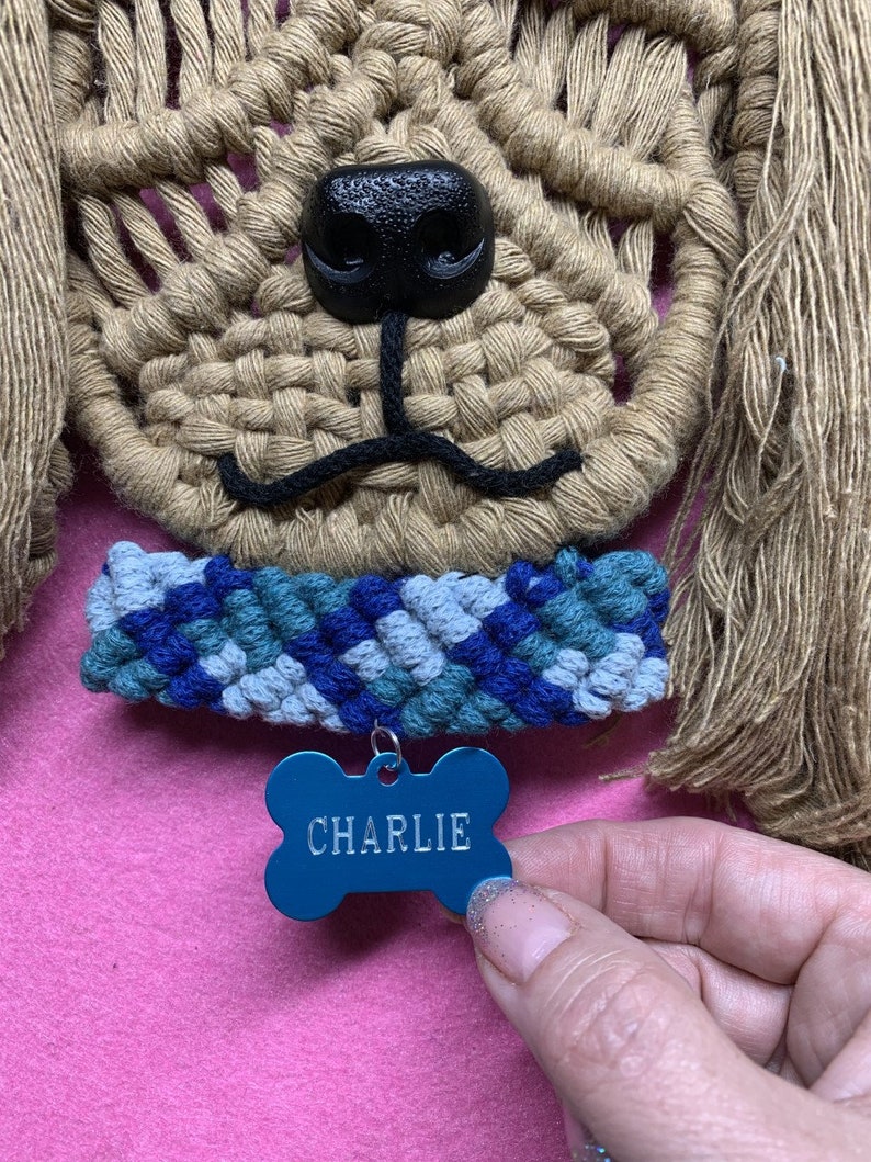 May include: A tan macrame dog wall hanging with a black nose and a blue and green macrame collar. The dog is wearing a blue bone-shaped tag that says "CHARLIE".