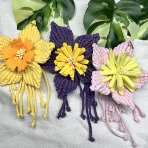 May include: Three macrame flower keychains with yellow felt flower centres. The keychains are in yellow, purple and pink.