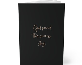 May include: Black notebook with the text "God penned this success story." in gold lettering.