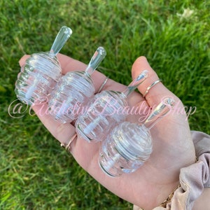 May include: Four clear, bee-hive shaped lip gloss containers with clear applicators are held in a hand. The containers are made of clear plastic and have a spiral design. The background is green grass.