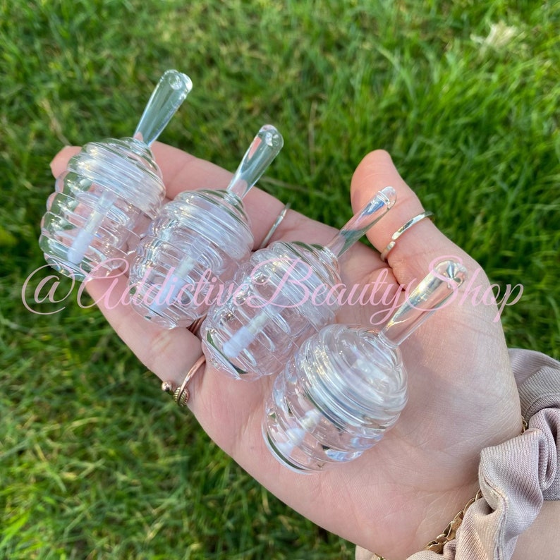 May include: Four clear, bee-hive shaped lip gloss containers with clear applicators are held in a hand. The containers are made of clear plastic and have a spiral design. The background is green grass.