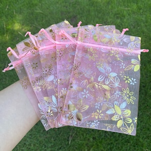 May include: A set of pink organza bags with a gold floral pattern. The bags are made of sheer fabric and have a drawstring closure.