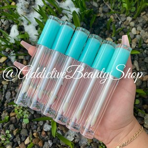 Puede incluir: Un grupo de ocho tubos de brillo de labios transparentes con tapas de color turquesa. Los tubos est&aacute;n dispuestos en forma de abanico y se sostienen en una mano. El texto "@Addictive Beauty Shop" es visible en la imagen.