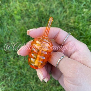 May include: An orange, honey-shaped lip gloss applicator. The applicator is held in a hand with manicured nails and silver rings. The background is blurred green grass. The text "@Addictive Beauty Shop" is visible in the image.