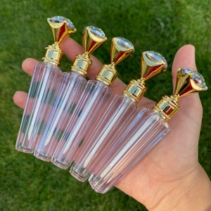 May include: Five clear lip gloss tubes with gold and diamond-shaped tops. The tubes are held in a hand.