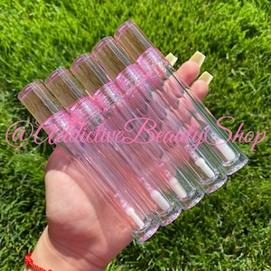 K&ouml;nnte beinhalten: Sechs transparente Lipgloss-R&ouml;hrchen mit rosa Deckeln und einem wei&szlig;en Applikator in Form eines Hirschfu&szlig;es. Die R&ouml;hrchen werden in einer Hand gehalten.